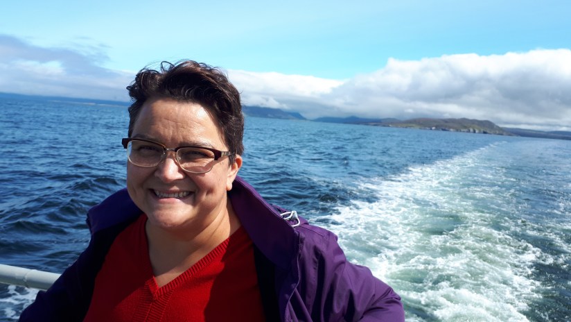 helga henry, smiling, standing with a railing at her back, the sea with a wake of a boat behind her.