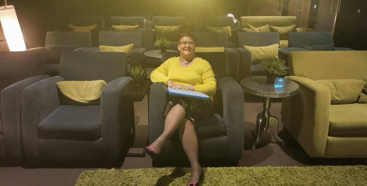helga henry, smiling, wearing a yellow jumper, sits in an empty auditorium with large armchair style seats in yellow and grey