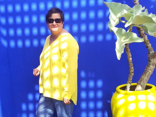 Woman in yellow top and blue trousers by a blue wall and yellow plant pot