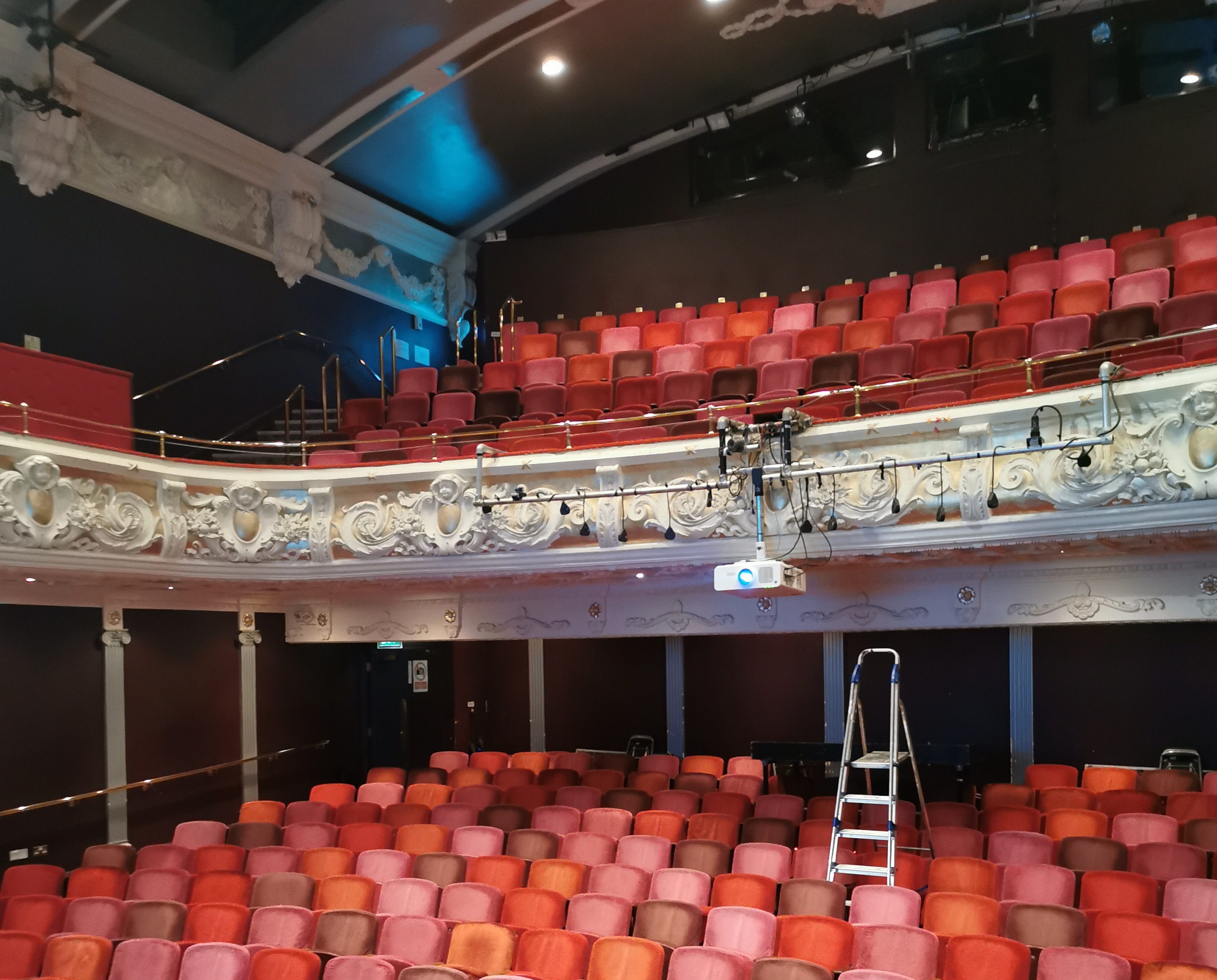 an empty theatre auditorium with rows of read seats and a balcony, a ladder in the centre of the room