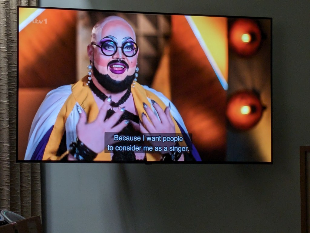 Television screen with a drag queen with a bald head, very heavy make up and silver nails.  Subtitle saying "Because I want people to consider me as a singer".